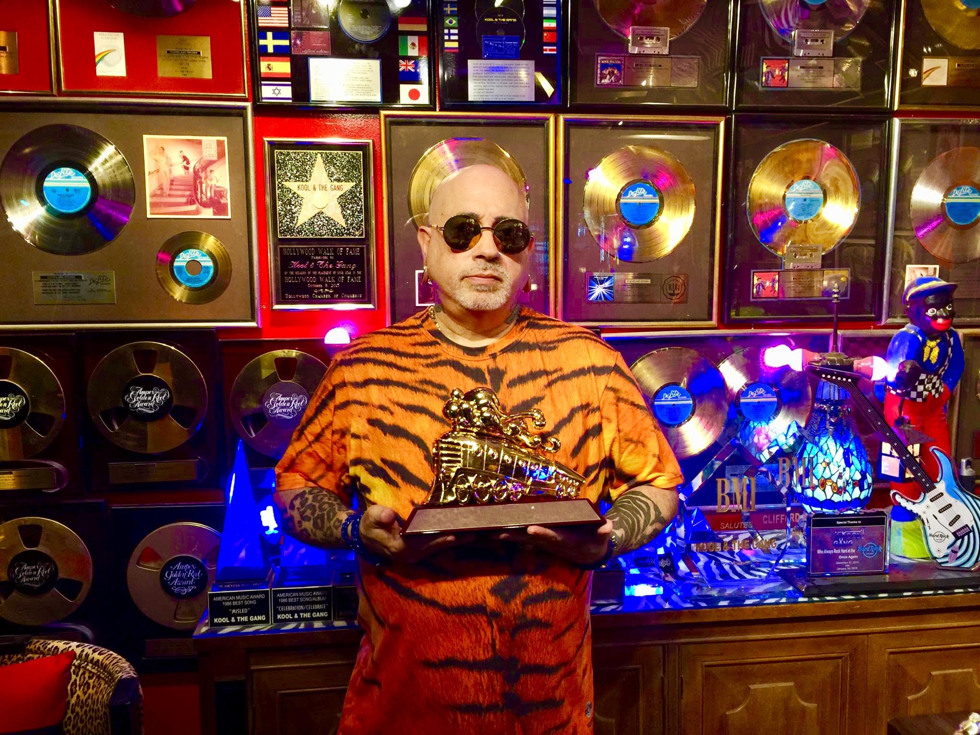 Cleveland 'Uhuru-Sasa' Brown holding the gold locomotive trophy before the Kool & The Gang gold-records wall